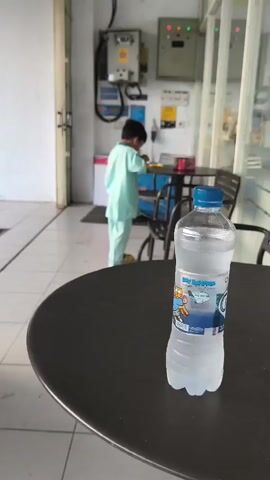 A bottle of water sits on a dark table in the foreground. In the background, a young child wearing a light blue outfit stands near some electrical equipment. The child is bent over and appears to be looking at something on the floor or a table. The scene appears to be indoors, possibly in a workshop or service area, with tiled flooring and some machinery visible.