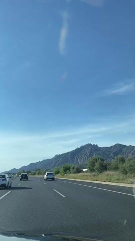 The video shows a view from a car driving on a highway, with mountains visible in the background. The sky is blue with light clouds. Several cars are visible on the highway. The road has multiple lanes and white painted lines dividing them. The car filming the video is in the rightmost lane. The scenery includes trees and other vegetation near the highway. The overall scene appears to be a sunny day during a road trip.