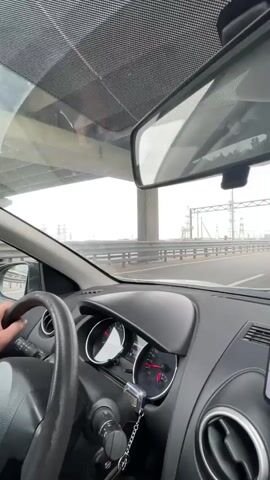 The video shows a view from inside a car driving on a highway. The driver's hands are visible on the steering wheel. Other cars can be seen on the highway, as well as high-voltage power lines on either side. The sky is overcast. The interior of the car is visible, including the dashboard, speedometer, and rear-view mirror.