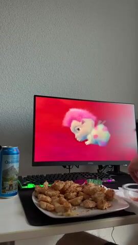 The video shows a person sitting at a desk with a computer monitor in front of them. On the monitor, a cartoon character is shown. In front of the monitor, on the desk, there is a plate of fried chicken, a can of soda, and a keyboard. A hand is visible on the right side of the frame, holding a fork and reaching towards the plate of chicken. The background is a plain, textured wall. The lighting suggests an indoor environment.