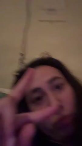 The video shows a person on a bed, possibly in a bedroom. The person is lying down and looking at the camera. The room is dimly lit, and there is a curtain in the background. There is also a teddy bear on the bed. The person is wearing a blue hoodie.