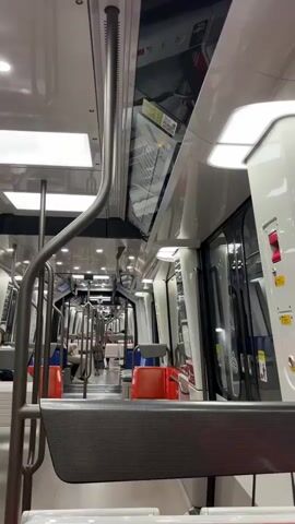 The video shows the interior of a nearly empty public transportation vehicle, likely a train or tram, moving. The camera is positioned looking down the length of the car. The seats are primarily red and blue with silver accents. The lighting is bright, coming from overhead panels. There are a few passengers visible sitting in the seats. A metal pole runs along the side of the aisle. The exterior of the vehicle is visible through the windows on the right, showing blurred scenery. A map or diagram is displayed on the ceiling in the distance. The overall impression is one of clean, modern public transit.