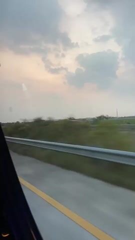 The media file depicts a view from inside a vehicle, likely a bus or car, looking out at a highway scene. The foreground is dominated by a portion of the road with a yellow dividing line and a metal guardrail running alongside it. Beyond the guardrail, a flat, grassy landscape stretches into the distance, dotted with scattered trees and some distant structures.

The sky above is a prominent feature, filled with soft, diffused clouds that suggest either early morning or late afternoon light. The overall lighting is muted and hazy, creating a serene and somewhat melancholic atmosphere. There are no people visible in the frame, nor are there any distinct objects that stand out as subjects.

The activity taking place is inferred to be travel, with the blur of the landscape indicating movement. The setting appears to be a rural or semi-rural area along a highway, given the open fields and the presence of road infrastructure.

Notable details include the cloudy sky, which hints at a possibly overcast or transitional weather condition. The time of day is likely around dawn or dusk due to the soft, diffused light. While the specific location is provided as Karangsari, Indonesia, there are no explicit visual cues within the frame to confirm this. There is no visible text in the media file. The emotional tone conveyed is one of peaceful transit, with the vastness of the sky and landscape evoking a sense of journey and contemplation.