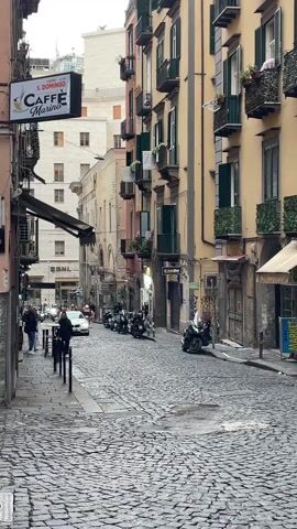 The camera faces down a narrow cobblestone street lined with buildings of varying heights and colors, primarily yellow and tan. Balconies with green shutters and flower boxes are visible on some of the buildings. On the left, a sign for "S. Domingo Caffe Marino" hangs above an awning. People walk along the sidewalk on the left. A white car drives slowly down the street, passing parked motorbikes. Other motorbikes are parked along the right side of the street. The sky is overcast.