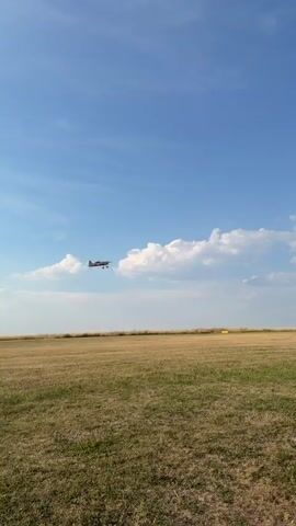 A small, red and white propeller airplane is captured in mid-flight against a bright blue sky with scattered white clouds. The airplane is positioned in the upper center of the frame and appears to be ascending. Below the airplane, a vast, dry, grassy field stretches to the horizon, indicating an outdoor, possibly rural, setting. The sun is shining, casting a bright light on the scene. The video seems to be shot from the ground, looking up at the aircraft.