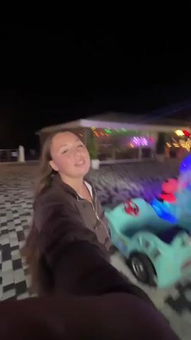 A woman wearing a brown hooded jacket and a white shirt is spinning in a circle in front of a brightly lit amusement park ride. The background is blurred due to the spinning motion, but hints of lights, buildings, and other rides can be seen. The ground is paved with a black and white patterned tile. The woman is looking at the camera and appears to be smiling and having fun.