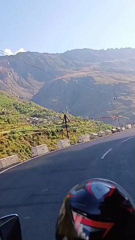 The video shows a first-person perspective of someone riding a motorcycle on a winding mountain road. The road is paved and has white lane markings. To the left of the road, there are steep mountain slopes covered in vegetation and some structures. To the right, a sheer rock face rises dramatically. The sky is clear and blue with some scattered clouds, and the sun is shining brightly, casting shadows on the road and mountains. The motorcycle rider is wearing a helmet, the front of which is visible in the foreground. The overall scene depicts a scenic and somewhat perilous journey through a mountainous region.