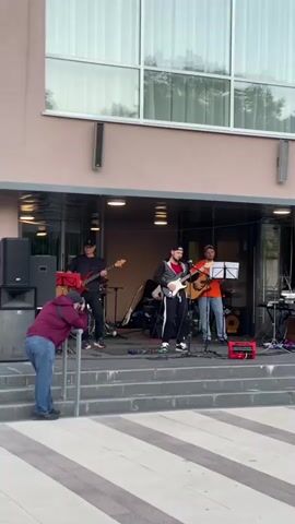 A street performance is taking place in front of a building. A band of three musicians is set up on a stage. The musician on the left is playing a bass guitar. The musician in the middle is playing an electric guitar and singing. The musician on the right is playing an acoustic guitar and singing. In front of the stage, a person is taking a photo of the band with a camera. The building appears to be a modern structure with large windows. The ground in front of the stage is paved with striped patterns. The lighting suggests it is daytime, possibly late afternoon or early evening.