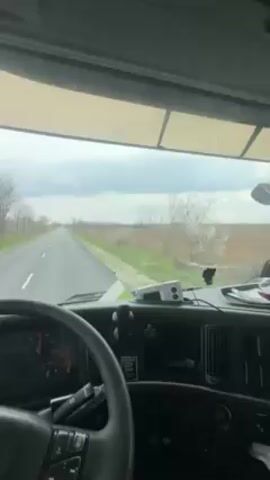 The video shows a view from inside a truck cab, driving down a straight, empty road. The driver's hands are visible on the steering wheel. The landscape outside consists of bare trees and fields. The side mirrors show a red truck following behind. The weather appears overcast. Music can be heard in the background.