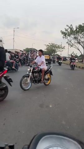 The video shows a street scene with many people riding motorcycles. The sky is overcast, suggesting it might be early morning or late afternoon. There are buildings and trees visible on the side of the road. Some of the motorcycles are carrying objects, such as cages stacked on top of each other. The people on the motorcycles are wearing casual clothing. The overall impression is a busy street with a lot of motorcycle traffic.