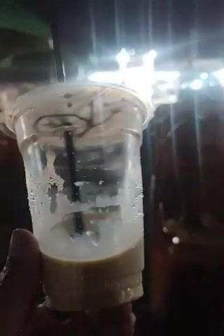 A person holds a plastic cup filled with coffee with a straw. The cup is partially empty, with a liquid layer at the bottom. Condensation is visible on the inside of the cup. The background is dark and out of focus, with bright lights in the distance. The person says, "This is the coffee. Coffee is amazing."