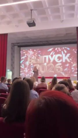 The video shows a young woman singing on a stage in front of a large screen. The screen displays the text "ВЫПУСК 2026" which translates to "GRADUATION 2026". The background of the screen is a vibrant display of fireworks. The woman is wearing a long, light brown dress and is holding a microphone. She appears to be the center of attention, with an audience seated in front of the stage. The audience members are mostly young people, likely students, and they are seated in rows of red chairs. Some audience members are visible from the back, with their hair styles and clothing suggesting a relatively casual event. There are some balloons visible on the stage, adding to the celebratory atmosphere. The lighting in the room is bright, suggesting it is daytime or a well-lit indoor venue. The overall scene conveys a sense of a graduation ceremony or celebration. The setting appears to be an auditorium or a large hall.