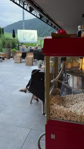 The video shows an outdoor seating area with a popcorn machine in the foreground. A large screen is visible in the background displaying a presentation. There are people seated in chairs, watching the screen. The environment appears to be a restaurant or a venue with an outdoor cinema. The overall atmosphere seems relaxed and casual.