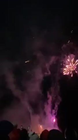 The video shows a fireworks display at night. The fireworks are various colors, including gold, red, and pink, and explode in the sky with a loud noise. There is a lot of smoke from the fireworks. In the foreground, there are a few people standing and watching the fireworks. Some are wearing hats, and some are holding red balloons. The sky is dark, and there are a few stars visible.