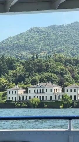 This media file captures a scenic view of a grand white villa situated along the shore of a large body of water, likely a lake, given the context of San Giovanni, Italy. The villa is a multi-part structure with a central, more imposing section flanked by two smaller, symmetrical wings. It features numerous arched windows and doors, suggesting a classical architectural style. Lush green trees and manicured bushes surround the villa, extending to the water's edge.

Behind the villa, a densely forested mountainside rises steeply, its slopes covered in vibrant green foliage. A thin, light-colored line, possibly a path or a historical funicular track, can be observed ascending the mountainside, adding a unique detail to the landscape.

The water in the foreground is a deep blue, rippling gently, suggesting a calm day. The sky above is a clear, bright blue with no visible clouds, indicating fair weather. The scene is viewed from a slightly elevated perspective, possibly from another boat or a higher vantage point on the shore, as indicated by the railing in the very bottom of the frame.

The time of day appears to be midday, with bright sunlight casting clear shadows and illuminating the scene. The overall impression is one of serene beauty and tranquility, highlighting the picturesque Italian landscape. There is no visible text in the media file.