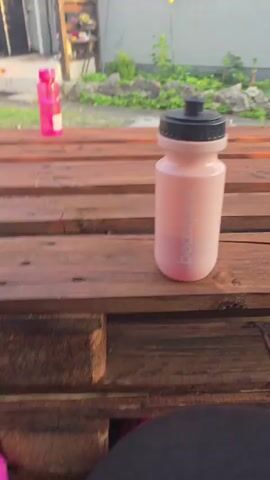The video presents a downward-facing shot from a first-person perspective, showcasing a person's feet, a wooden surface, and two water bottles in an outdoor setting.

In the foreground, the person's feet are clad in bright pink Crocs with light blue straps and white socks, with black pants or leggings partially visible. The Crocs are personalized with several charms, including a gold-colored chain spelling "love" on the left foot, and various sparkly, jewel-like embellishments on both. The feet are positioned beneath a rustic wooden structure, which appears to be constructed from stacked pallets or rough-hewn planks.

On this wooden surface, two water bottles are visible. To the right and closer to the camera is a frosted pink water bottle with a black lid, displaying the brand name "beandpink" printed vertically on its side. Further back and to the left, a smaller, clear pink water bottle with a white lid is placed.

The background reveals an outdoor environment with greenery, suggesting a garden or yard, and a building or structure in the distance. The lighting indicates it is daytime, possibly early morning or late afternoon. The overall scene conveys a casual and relaxed atmosphere. The person's feet shuffle slightly throughout the short video. The location context provided for this scene is Zirndorf, Germany.