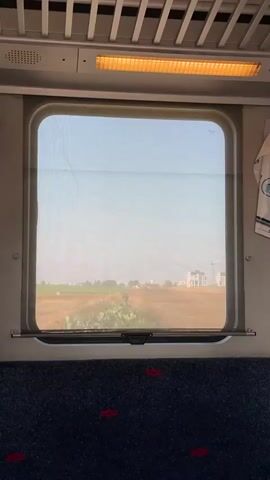 The video captures a view from inside a train, looking out a rectangular window with rounded corners. A translucent blind is partially lowered, giving the exterior scene a slightly diffused appearance. Inside the train, a dark blue patterned seat with red accents is visible in the foreground. Above the window, part of the train's ceiling and an orange-glowing overhead light fixture are seen.

Through the window, a bright, clear sky dominates the upper portion of the view, suggesting a daytime scene with sunny weather. The landscape outside consists primarily of wide, dry, light brown fields. As the train moves, various elements pass by: initially, a tall, slender, dark green tree, followed by clusters of distant white buildings, some appearing residential and others possibly under construction with visible cranes. Later, a road or path with green trees and bushes comes into view, along with more scattered buildings on the horizon. The changing scenery indicates the train is in motion, passing through a semi-rural or suburban area, consistent with locations such as Hod HaSharon, Israel. There are minor imperfections or cracks visible on the window glass, particularly in the upper left. A white piece of cloth or an item with an indistinguishable blue logo hangs to the right of the window.