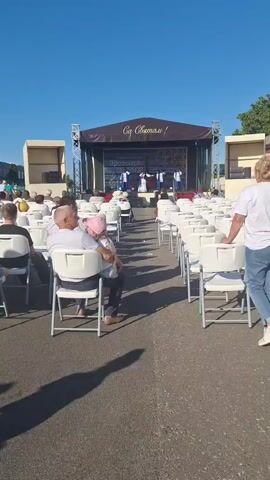 An outdoor performance stage is set up with a banner above reading "С праздником!". A group of performers, dressed in white costumes, are on stage. Rows of white chairs are set up in front of the stage, facing the performance area. Many audience members are seated in the chairs, some are standing. The sky is clear and blue, indicating it is daytime. In the foreground, a man is holding a young child, and a woman is walking away from the camera. Another woman is standing by a chair. There are shadows cast on the ground, suggesting the sun is high in the sky.