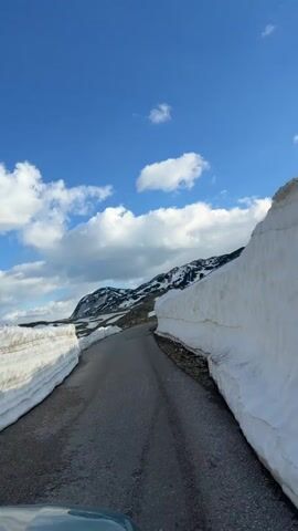 The video shows a road cut through thick snowbanks. The road is asphalt and appears to be well-maintained. The snowbanks on either side of the road are tall, reaching at least 6 feet in height. The snow is mostly white, with some dirt and debris visible. The sky is blue with scattered white clouds. In the distance, there are snow-capped mountains. The camera is positioned from a low angle, likely from the dashboard of a car. The car appears to be moving forward, giving the viewer the perspective of driving down the road.