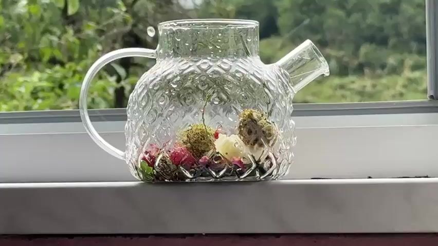 A clear glass teapot sits on a windowsill. The teapot contains dried flowers and herbs. Hot water is being poured into the teapot, creating steam. Outside the window, green foliage is visible.