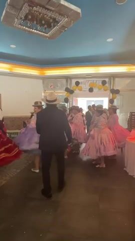 A man in a black suit and white hat dances in front of a group of women in layered, colorful dresses and hats. The women appear to be dancing in formation. The floor is dark tile. In the background, there is a doorway decorated with black and gold balloons. Above the doorway, there is a sign with writing in what appears to be Spanish. The room has a blue ceiling with decorative lighting fixtures. There are tables draped with white tablecloths on the right side of the room. The lighting suggests an indoor event.