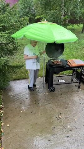 A person is grilling meat under a bright green umbrella in the rain. The umbrella is open and positioned over the grill to keep the rain off the food and the person. The person, wearing a white t-shirt and grey pants, is holding a pair of tongs and appears to be tending to the meat on the grill. The grill is a black, wheeled charcoal grill with its lid open. Several pieces of meat are cooking on the grates, and there is smoke rising from them. The ground around the grill is wet and speckled with fallen leaves, indicating that it is raining. Lush green foliage is visible in the background, suggesting an outdoor setting, possibly a backyard or park. The overall scene depicts a determined effort to grill outdoors despite inclement weather.