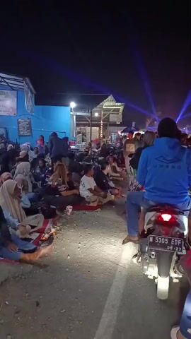 This is a vertical, low-light video taken at night from a slightly elevated perspective, looking down and across a street or open area where a crowd of people are gathered. The ground is paved with some loose gravel and dirt. Many people are sitting on the ground, some on mats or blankets. They are facing in various directions, with many appearing to be looking at something off-camera to the right. The crowd is diverse, with men and women, some wearing headscarves. In the foreground, a person on a motorcycle is parked, facing away from the camera and towards the crowd. The motorcycle's brake light is illuminated. In the background, there are buildings with lights on and structures that appear to be stalls or vendor booths. Purple and white spotlights are visible in the sky, suggesting an event or performance is taking place. The overall atmosphere appears to be a casual, outdoor gathering.