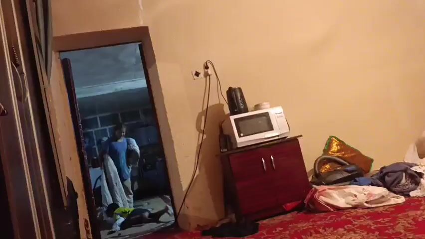 The video begins with a view of a cluttered room. In the foreground, to the left, is a partially open closet or cabinet with stacks of colorful blankets or textiles. The doorway to another room is visible in the center of the frame, angled to the left. Through the doorway, several people are seen. One person in a blue shirt and light-colored pants appears to be standing, while another person in a yellow vest is lying on the floor. A third person, wearing a light blue shirt and dark pants, is also visible in the doorway. The lighting suggests it could be daytime or artificial light within the room.

The main room features a light-colored wall. To the right, on a small, dark red cabinet, sits a white microwave oven and a black electric kettle. The cabinet has two doors with silver handles. To the right of the cabinet, there are various items on the floor, including what appears to be a large, shiny gold bag or fabric, a vacuum cleaner, and some bundled clothing. The floor is covered with a red rug with a patterned design.

The activity in the doorway appears to be a chaotic or potentially concerning situation, with one person on the floor. The overall impression of the scene is one of domestic disarray. There is no visible text in the video. The setting is identified as Naray, Kyrgyzstan.