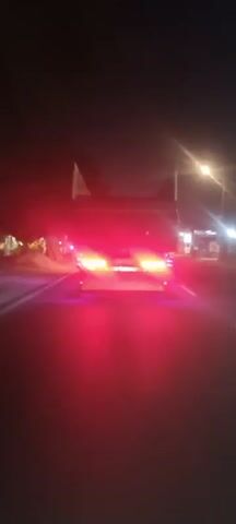 A video taken from the perspective of a moving vehicle at night shows the rear of a truck driving on a road. The truck has multiple red taillights that are illuminated. Streetlights and buildings are visible on either side of the road. The camera is shaking slightly, suggesting the vehicle is in motion.