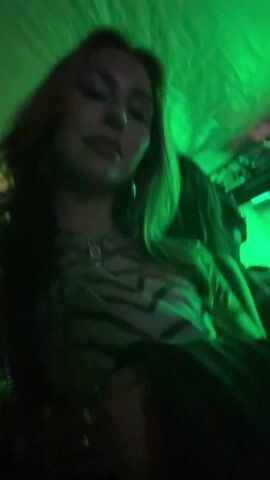 A woman is recording herself at a crowded indoor concert. The venue is lit with red and green lights and smoke. The woman appears to be smiling and enjoying the music. The crowd is dancing and jumping.