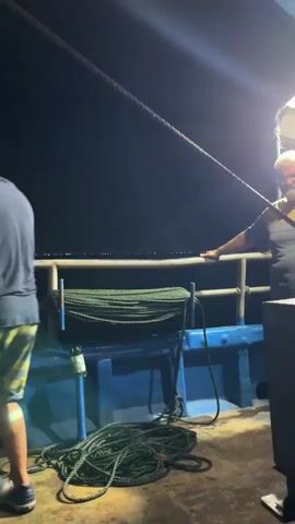 The video shows a person on a boat at night, holding onto a rope. A large fishing net is being lowered from above. The boat appears to be on the water, with the dark sea and a distant light visible in the background. Another person's arm is visible on the right side of the frame, also appearing to be on the boat. There is a large spool of green rope on the boat, as well as other coiled ropes. The net is round and appears to be made of mesh material. The overall impression is of a fishing operation taking place at night.