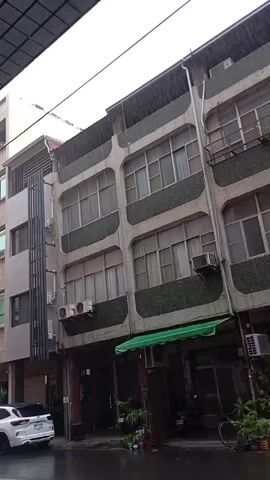 The video captures a rainy street scene in an urban area, identified as Kaohsiung, Taiwan. The footage begins by showing the upper floors of several multi-story buildings, then pans down to reveal the street level, where heavy rain is visibly falling.

The scene features a row of multi-story buildings, likely residential or mixed-use. Many of these structures exhibit a distinctive architectural style with rounded balcony fronts and textured, greenish-grey mosaic-like tiling. Numerous air conditioning units are mounted on the exterior walls. The ground floors include various facades, some with roll-down shutters and others with glass doors, suggesting a mix of residential entrances, garages, or small businesses. A green awning is extended over one of the ground-floor units.

The street is visibly wet from the continuous downpour, with raindrops clearly impacting the asphalt surface and creating splashes. A yellow line is painted on the wet road.

Vehicles present in the scene include a white Ford SUV parked on the left side of the street, displaying a partial license plate. Several scooters or motorcycles are parked along the sidewalks in front of the buildings, and a bicycle leans against a wall. Potted plants are arranged near the entrances of some buildings, adding small touches of greenery. The sky above is overcast, indicative of the rainy weather. No people are visible in the video. The only discernible text is the "FORD" logo on the white SUV and parts of its license plate.