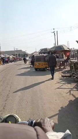 The video shows a street scene in what appears to be a busy market area. The camera is mounted on a motorcycle, providing a rider's point-of-view. The road is paved and lined with motorcycles parked on both sides. People are seen standing and walking around the motorcycles and small shops. A yellow tricycle taxi is also visible. Overhead are power lines and the sky is clear. The rider slowly drives forward, navigating through the crowd of people and parked motorcycles.