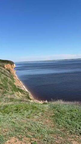 The video shows a wide, dark body of water under a bright blue, cloudless sky. The camera pans slightly to the right, revealing a long barge being pushed by a tugboat in the distance. The foreground features a grassy cliff edge.