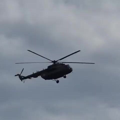The video shows a dark-colored helicopter in mid-flight against a cloudy sky. The helicopter is silhouetted and its rotors are blurred due to motion. It appears to be a military or transport helicopter, with landing gear visible. The setting is likely outdoors, with the sky indicating it's daytime. The clouds suggest either overcast or partly cloudy weather. There are no people visible. No specific location cues are evident from the sky or helicopter itself, other than the user-provided context of "Новостройка, Russia". The overall mood is neutral, focusing on the mechanics of the aircraft in its environment. No visible text is present on the helicopter or in the sky.