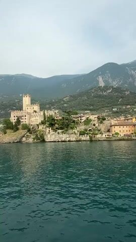 The media file is a video showing a panoramic view of a historic castle on a rocky promontory overlooking a large body of water, likely a lake. The castle, made of light-colored stone, has a prominent tower and appears to be part of a larger complex with surrounding buildings and greenery. A town or village with red-tiled roofs is visible along the shoreline to the right of the castle. In the background, steep, tree-covered mountains rise up, with a hazy, possibly cable car track visible on one of the slopes. The water in the foreground is a deep teal color with visible ripples, suggesting it's a calm day. The sky is overcast, indicating possibly early morning or late afternoon, or generally cloudy weather. A small boat is visible on the water to the left of the castle. The scene evokes a sense of historical grandeur and natural beauty, with the imposing castle set against the dramatic mountainous backdrop. The location is identified as Malcesine, Italy, which is known for its Scaligero Castle situated on Lake Garda. There is no visible text within the media file.
