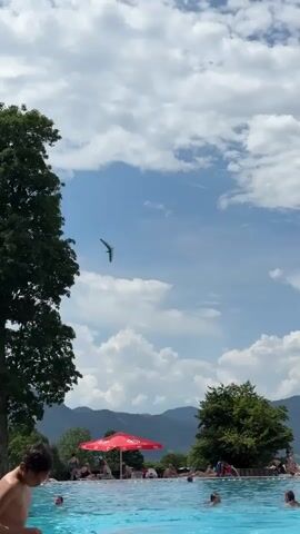 A drone is flying in the sky above a swimming pool. Several people are in the pool, and some are sitting by the edge. There are trees and mountains in the background. One person jumps into the pool, creating a large splash.