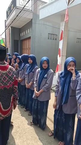 A group of young women in blue hijabs and grey tops stand in a line. They are dressed in matching outfits with blue patterned skirts. A man in a black cap and a patterned red and black shirt stands facing them. The women appear to be listening or observing. The background shows a building with a dark grey facade and a white door. There are strings of small red and white flags hanging across the street, suggesting a celebratory or festive occasion. The ground is paved with light-colored stones.