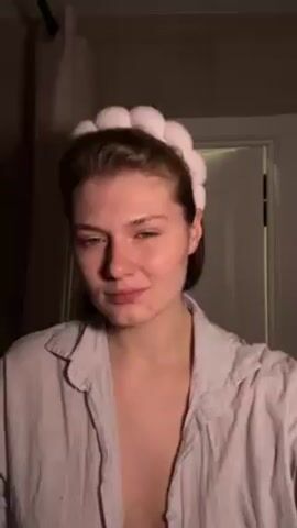 A woman with short brown hair wearing a light-colored striped shirt and a white fluffy headband is centered in the frame. She is in a dimly lit room, possibly a bathroom, as a dark curtain rod and a closed white door are visible in the background. The woman is applying a liquid to her face with her hands, rubbing it in a circular motion. Her fingernails are painted dark red. She is looking at the camera while performing this action.
