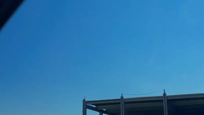 A clear blue sky is visible with a few thin contrails. On the right side of the frame, the top of a modern building under construction or with an open roof structure is visible, supported by steel beams. On the left side, a tall, white, cylindrical pole, possibly a utility pole or part of a larger structure, is present. Trees are visible in the distance on the right horizon. The camera appears to be moving, likely from a vehicle.