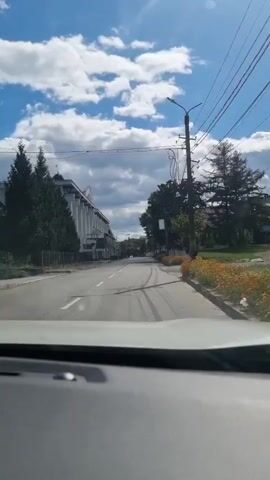 The video begins from the perspective of a driver looking out from inside a vehicle. The road ahead is visible, with a building on the left and trees and other structures on the right. The sky is blue with scattered white clouds. As the vehicle moves forward, the building on the left appears to be a large, multi-story structure with a white facade and columns. The road has lane markings and some tire tracks. On the right side, there are trees, a utility pole with wires, and some residential buildings. The vehicle passes by a crosswalk with red and white stripes. The overall impression is of a drive through a suburban or semi-urban area on a sunny day.