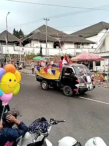 The video shows a street scene with a procession or parade. A small black pickup truck is decorated with flags and banners and is moving down the street. Several people are riding in the back of the truck, some holding Indonesian flags. In the foreground, a person is holding a bunch of colorful balloons, including one shaped like a smiley face. Other people are walking along the side of the street, and there appear to be stalls or vendors set up. Buildings with red and white striped flags are visible in the background, suggesting a celebration, possibly Independence Day. The sky is overcast. There are also motorcycles parked in the foreground.