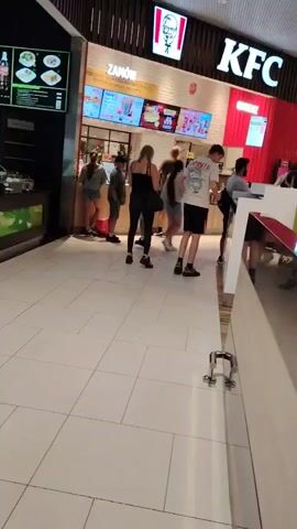This media file shows an indoor scene, likely a food court or a shopping mall, in Wołomin, Poland. The time appears to be daytime, given the lighting.

The primary subjects are several people, including adults and children, standing in a queue. They are facing a food counter, suggesting they are waiting to order or pick up food. One child, a young girl with blonde hair in a ponytail, is walking away from the counter towards the camera, wearing a pink t-shirt and a brown skirt. Other individuals in the queue are dressed casually.

Prominently visible are the logos and signage for KFC, a popular fast-food chain, indicating a KFC outlet is present. Above the counter, digital menu boards display food items and prices. Another sign with text, likely the name of a food establishment or a special offer, is visible above the menu boards. The flooring is tiled with a light-colored square pattern. The setting is clean and well-lit, typical of an indoor public space.

The activity taking place is people queuing at a food establishment. There are no explicit indicators of emotions, but the general atmosphere appears to be neutral, suggesting a routine activity like dining or shopping. The presence of multiple people and the food counter suggest a moderately busy period.