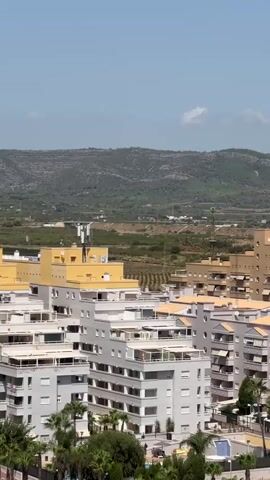An aerial perspective captures a cityscape with several multi-story buildings, some painted yellow, others in various shades of beige and white, with balconies and awnings. Palm trees are visible in the foreground. In the background, rolling green hills under a clear blue sky with a few wispy clouds form the horizon. The scene is likely a sunny day in a Mediterranean or similar climate.