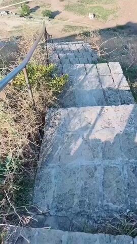 This video shows a person's point-of-view descending a set of weathered concrete stairs. The stairs appear to be outdoors, with dry brush and some green foliage growing on either side. A metal handrail is visible along the left side of the stairs. In the distance, a landscape with buildings and a dirt road can be seen. The lighting suggests it is daytime, possibly late afternoon due to the shadows. The camera movement indicates the person is actively walking down the stairs.