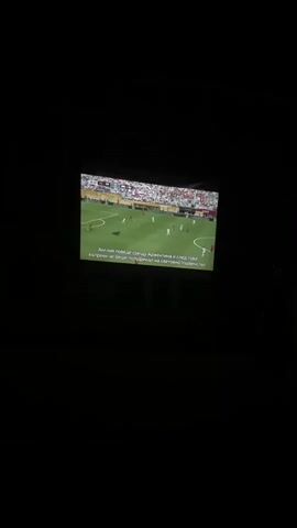 A dimly lit room with a projected image of a soccer game on a screen. The game appears to be a professional match with players in red and white uniforms on a green field. There are spectators in the stands behind the players. Text overlays the bottom of the projected image, indicating that this is a broadcast of a soccer game with subtitles. The scene is dark, suggesting the room is not well-lit, and the projection is the primary light source.