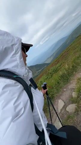 The video shows a person hiking in the mountains, captured from a first-person perspective. The hiker is wearing a white hooded jacket and sunglasses, and they are holding two trekking poles. The scene is a mountain slope with a trail. The background features a scenic view of mountains and a cloudy sky. The hiker is looking out at the landscape and adjusting their grip on the trekking poles.