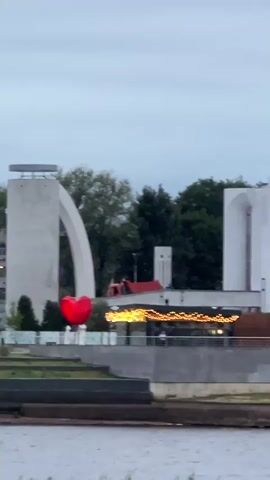 The video shows an outdoor scene with a large red heart-shaped balloon suspended in the air. The balloon is positioned in front of a white architectural structure with a large arch. In the background, there are trees, buildings, and a body of water. A restaurant or cafe with string lights is visible on a lower level. There are a few people walking on the patio of the establishment. The sky is overcast, suggesting it is either dawn or dusk. The water in the foreground appears choppy.