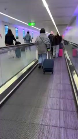 The video shows a group of people on a moving walkway in what appears to be an airport. They are walking in the same direction, and most of them are pulling luggage behind them. The walkway has a textured surface and is bordered by glass railings on either side. The environment has bright overhead lighting and colorful geometric designs on the walls. One person walking on the walkway is wearing slippers or slides.