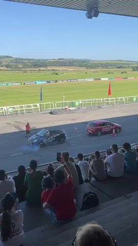 A black car performs a donut on a paved surface, emitting smoke from its tires. A red car is parked nearby. A person in an orange vest stands to the left of the black car, appearing to direct the action. In the foreground, a crowd of spectators sits in tiered seating, watching the event. Behind the paved area is a large, green field with white fencing and various flags. The sky is clear and blue. The overall scene appears to be an outdoor event, possibly a motorsport demonstration.